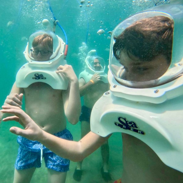 Three people underwater wearing helmets with air tubes, swimming in clear water.