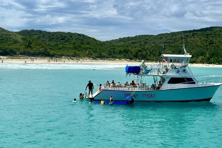 Boat with people in turquoise water near a sandy beach and green hills.