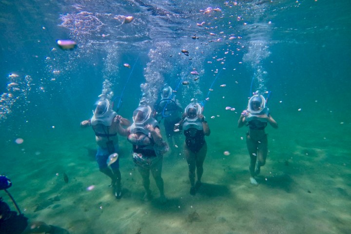 Group of people walking underwater with helmets, bubbles around.