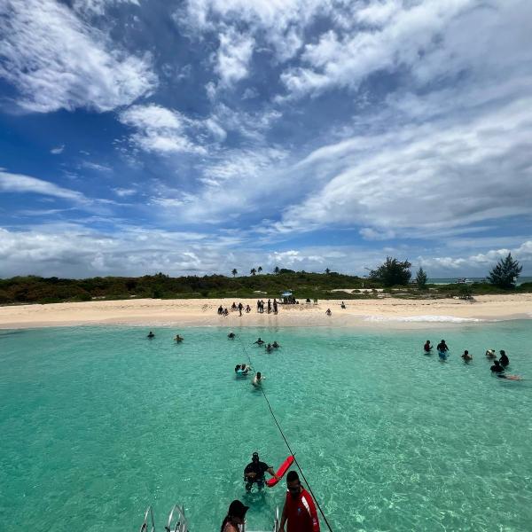 People swimming in clear turquoise water near a sandy beach under a cloudy sky.