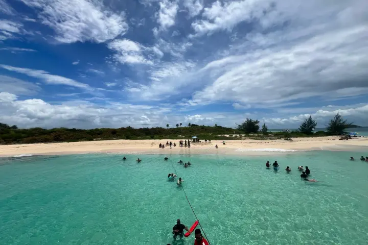 People swimming in clear turquoise water near a sandy beach under a cloudy sky.