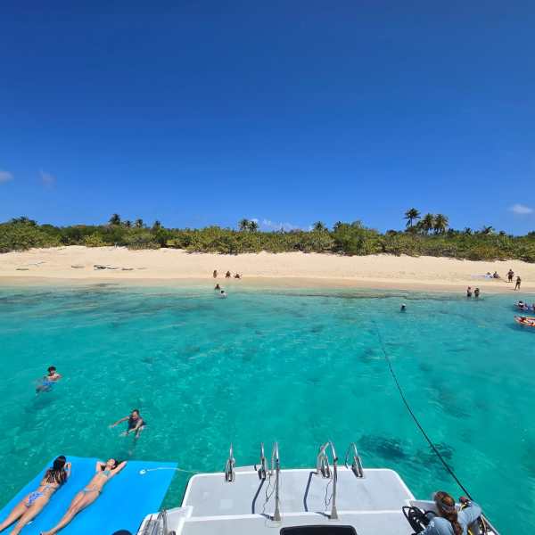 People relax on a boat near a beach with clear blue water and a sandy shoreline.