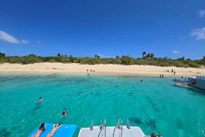 People relax on a boat near a beach with clear blue water and a sandy shoreline.