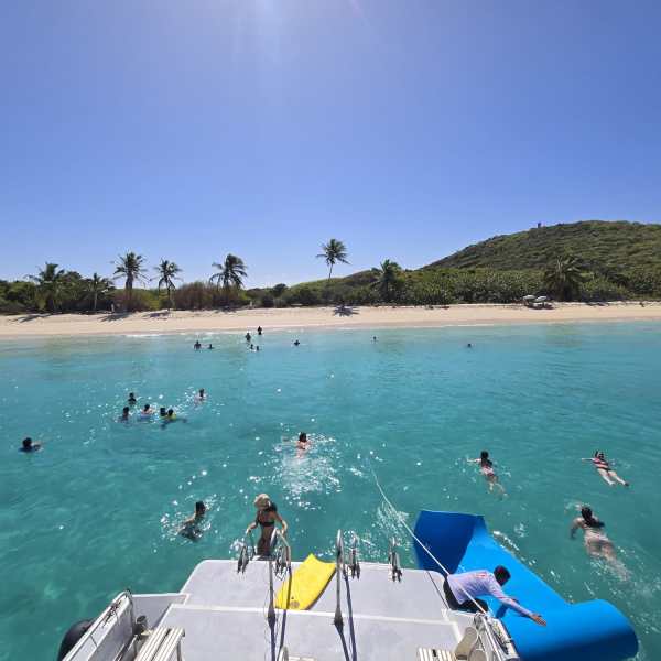 View from boat of swimmers in clear turquoise water near a sandy beach with palm trees.